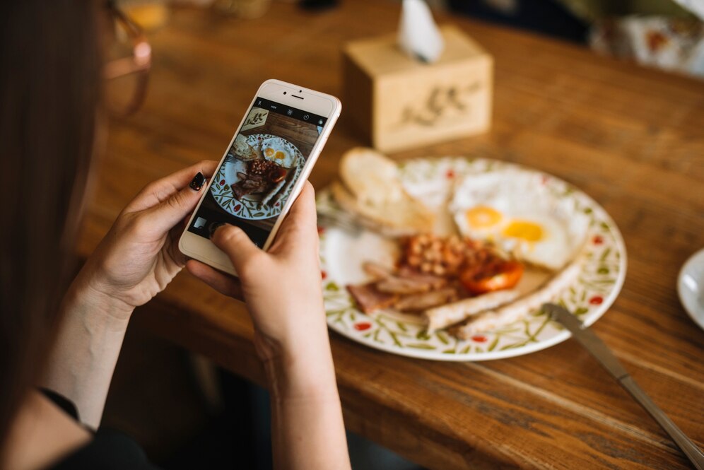 Hands holding smartphone to photograph breakfast plate with eggs, beans, and toast on wooden table