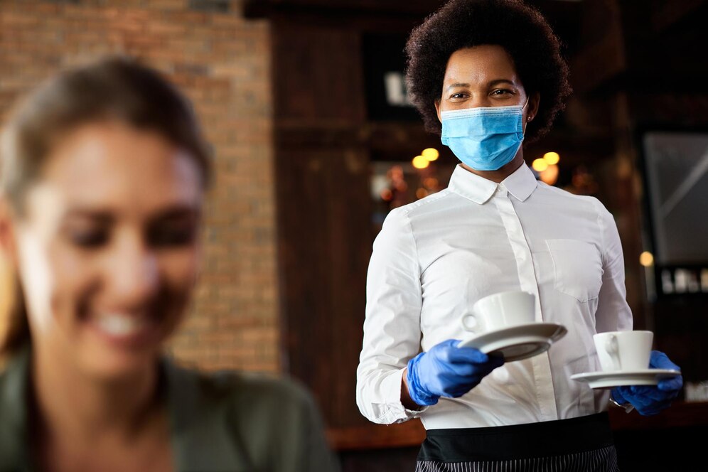 Restaurant staff in masks and gloves serving coffee in a restaurant setting to a smiling woman in the foreground